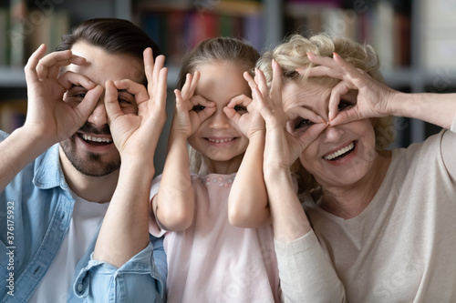 Head shot portrait smiling little girl with father and grandmother making funny faces, binoculars glasses eyewear shape gesture, overjoyed mature woman with son and granddaughter looking at camera