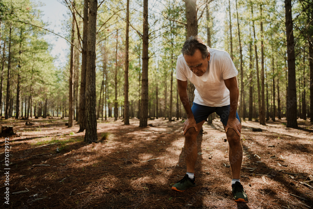Fit elderly male tired bending over with hands on knees in green forest. Fit elderly male tired bending over with hands on knees in green forest.