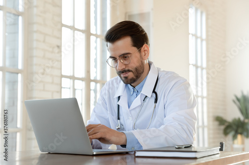 Focused young doctor wearing glasses working on laptop in office, sitting at desk, looking at screen, serious therapist gp writing report or email, consulting patient online, filling medical card