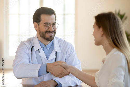 Close up smiling doctor therapist gp wearing white medical uniform and glasses shaking patient hand at meeting in hospital, getting acquaintance, making healthcare insurance deal, agreement