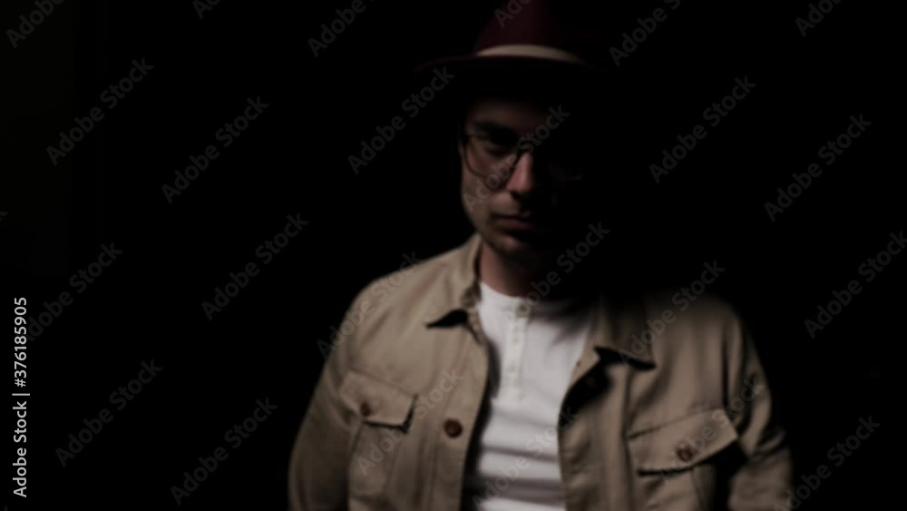 young fashion model in beige jacket showing from the dark, arranging glasses, looking up, sensually touching neck, adjusting hat, moving and fixing jacket, walking in a shadow on black background young fashion model in beige jacket showing from the dark, arranging glasses, looking up, sensually touching neck, adjusting hat, moving and fixing jacket, walking in a shadow on black background