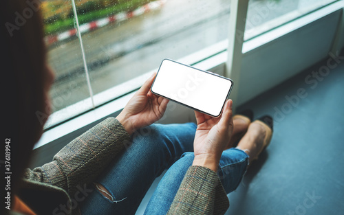 Mockup image of a woman holding  mobile phone with blank white desktop screen while sitting on the floor