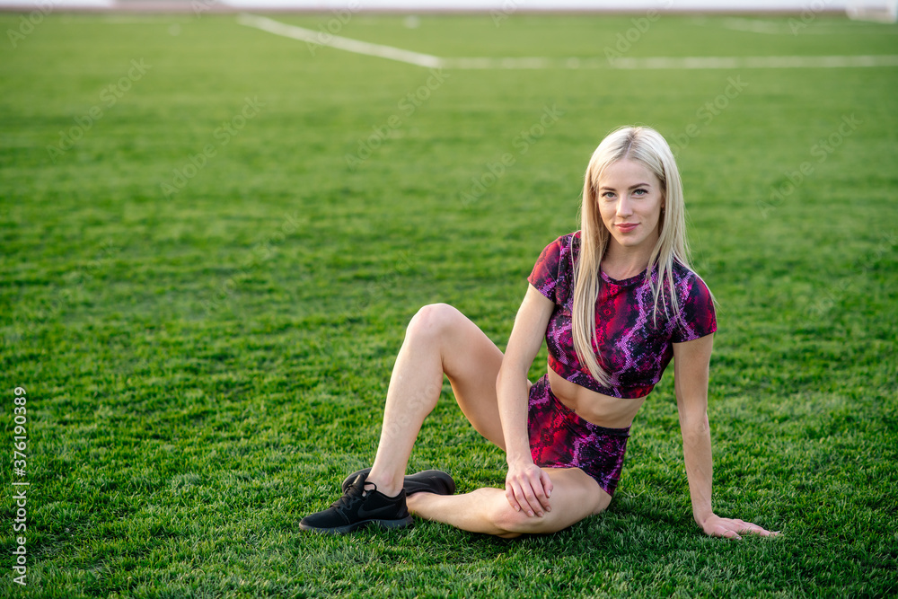 Beautiful young blonde woman in sportswear sits on the grass in the stadium before training