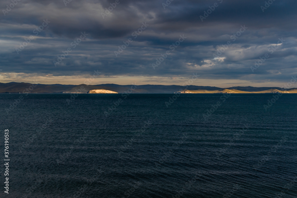 view of the clear calm undulating dark blue water of Lake Baikal, gold mountains in sunset light on the horizon
