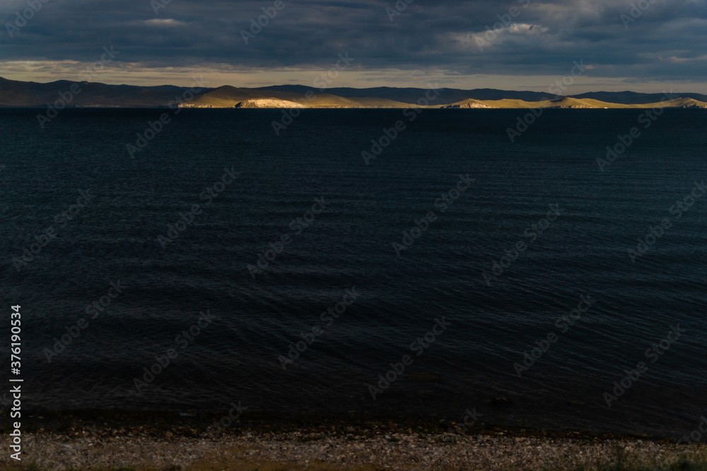 Fototapeta premium view of the clear calm undulating blue water of Lake Baikal, sunset mountains on the horizon, clouds, coast
