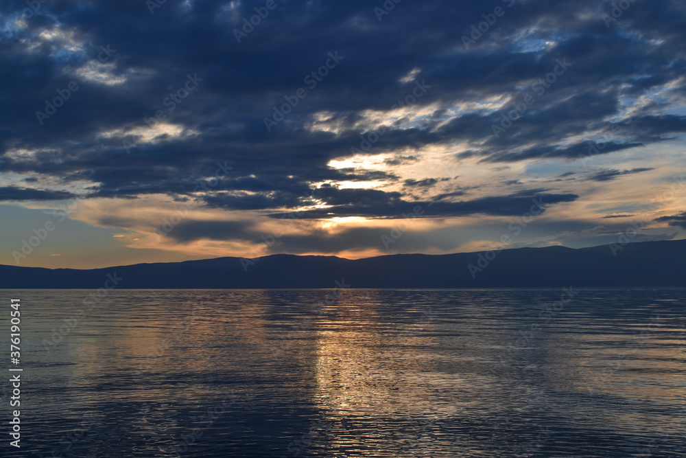 view of the clear calm undulating blue water of Lake Baikal, mountains on the horizon, sunset sky, clouds, reflection