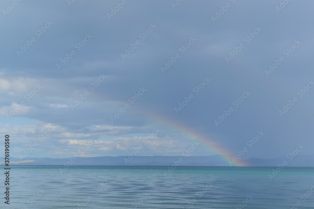 view of the clear calm undulating blue water of Lake Baikal, mountains on the horizon, colorful rainbow after rain