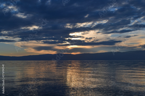 view of the clear calm undulating blue water of Lake Baikal, mountains on the horizon, sunset sky, clouds, reflection