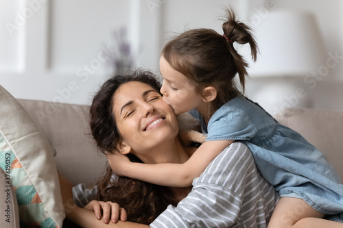 Little daughter gently cuddles kiss mother on cheek showing love and express caress resting on couch at home. Happy family, pleasure be mommy, mother day congratulations, sweet moment together concept