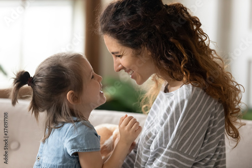Fotografi Side view young beautiful mother and little pretty daughter sit on couch at home smile looking at each other with tender and love