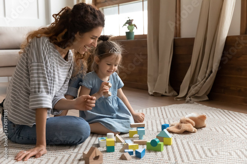 Mom and little daughter play with color wooden bricks toys set. Baby sitter and small kid girl sit on warm floor on carpet enjoy funny game together. Educational activity, development of child concept