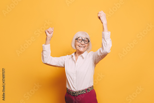 Happy elderly woman with white hair, round glasses wearing white blouse, red pants and leopard print belt standing isolated over orange background with raised arms.