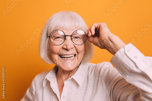?lose-up senior woman with blonde short hair wearing light blouse, smiling and fixing her glasses by her hand. Woman isolated over bright orange background.