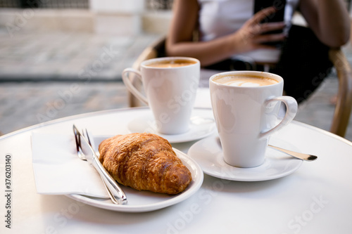 Two lattes coffee in white cups on a saucer and a croissant in a white plate  a fork and a knife on a summer terrace in a cafe. Girl with a phone in the background.