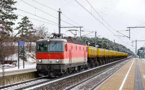 A large, powerful electric locomotive in red and white is pulling a train of freight cars. Passenger platform. Winter snowy weather.