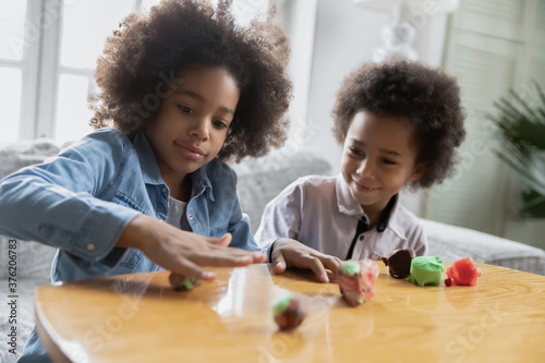 Happy african american older sister and younger brother involved in creating figures with plasticine at home, interested little children siblings developing creativity, improving fine motor skills.