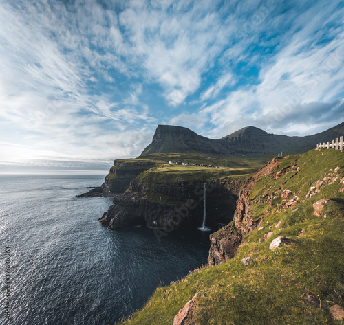 Gasadalur village and Mulafossur its iconic waterfall during summer with bluw sky. Vagar, Faroe Islands, Denmark. Rough see in the north atlantic ocean.