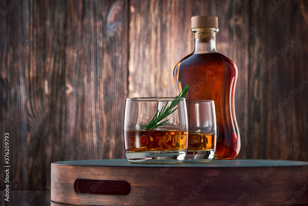 Two round glasses of whiskey with ice and a sprig of rosemary, and a full bottle of whiskey, stand on a tray against an old wooden wall. Two round glasses of whiskey with ice and a sprig of rosemary, and a full bottle of whiskey, stand on a tray against an old wooden wall.