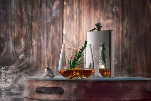 A crystal glass of whiskey with a sprig of rosemary and a metal flask, shrouded in clouds of smoke, stand on a tray against an old wooden wall.