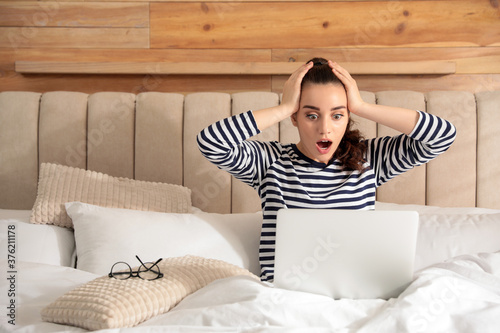Shocked young woman working with laptop on bed at home