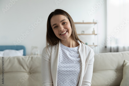 Happy young girl sitting on sofa indoors, looking at camera, enjoying video call with friends. Professional female blogger streaming in social network online, talking to followers webcam view.