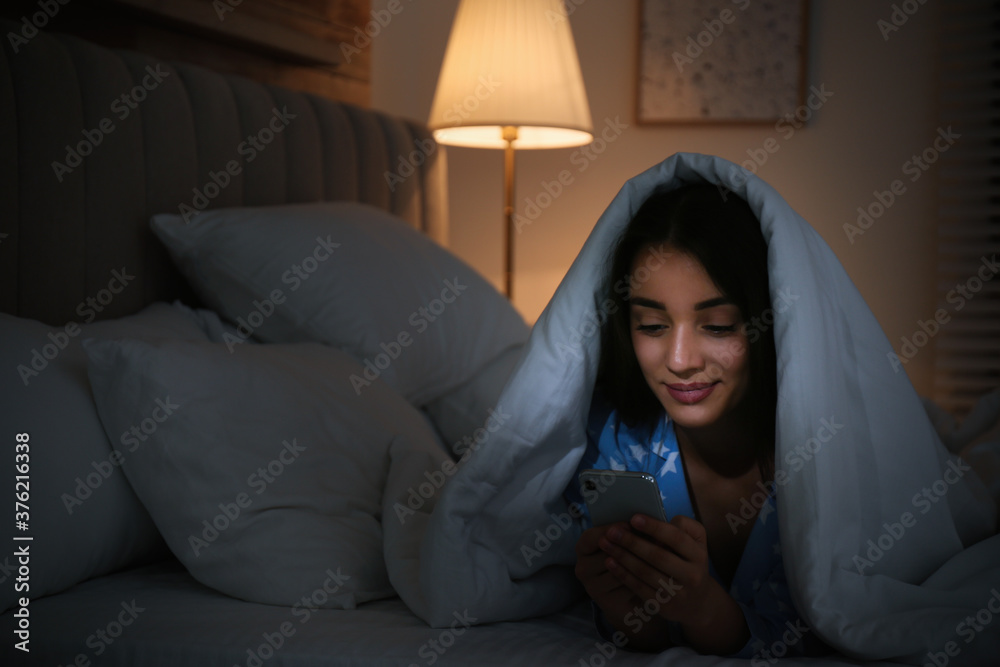 Happy woman using mobile phone under blanket in dark bedroom Happy woman using mobile phone under blanket in dark bedroom