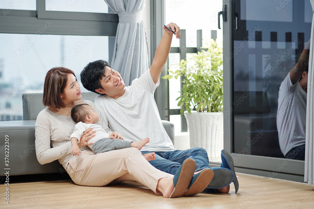 Smiling handsome Asian young man taking selfie with his wife and little daughter when they are sitting on the floor Smiling handsome Asian young man taking selfie with his wife and little daughter when they are sitting on the floor