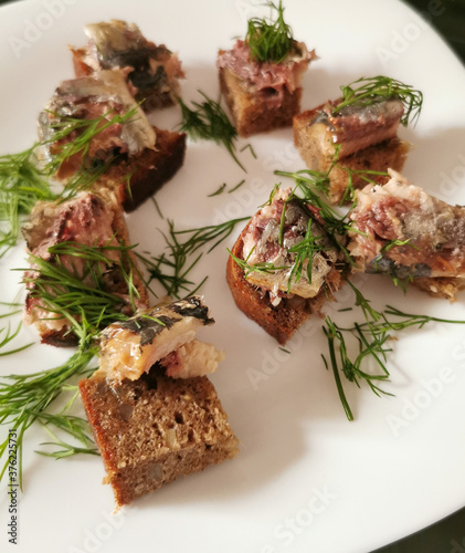 black rue bread and pieces of smoked sardines sprinkled with dill on a white plate, snacks to the buffet