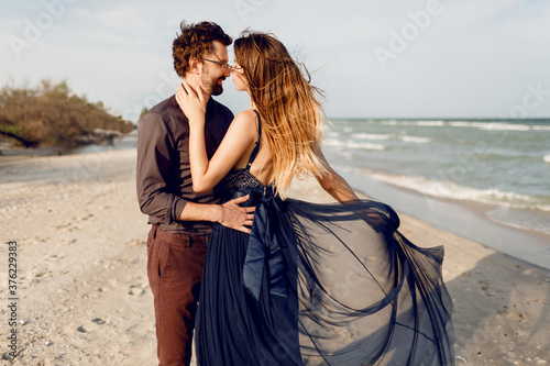 Romantic moments of beautiful couple, fashionable woman and man posing outdoor near the sea. Amazing blue dress and casual outfit. Honeymoon vacation.