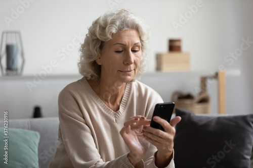 Focused mature woman using phone at home, looking at screen, interested middle aged senior female holding smartphone, chatting online, browsing mobile device apps, sitting on couch