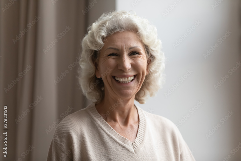 Head shot portrait smiling mature woman with grey curly hair standing at home, beautiful overjoyed middle aged senior female looking at camera, feeling positive, posing for photo, happy retirement