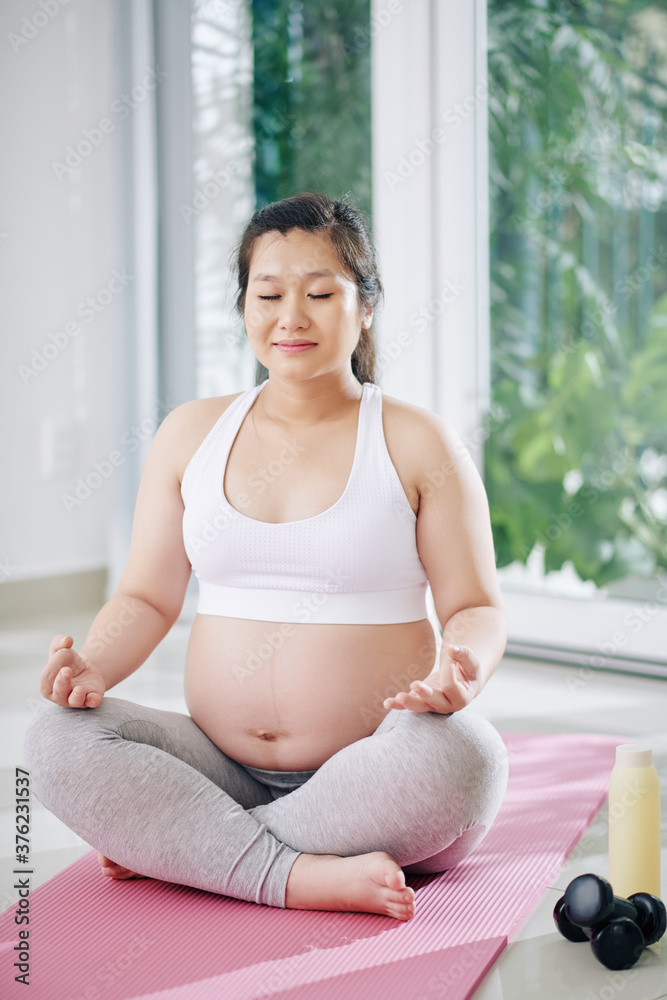 Smiling young pregnant Vietnamese woman sitting in lotus position, closing eyes and doing breathing exercise Smiling young pregnant Vietnamese woman sitting in lotus position, closing eyes and doing breathing exercise
