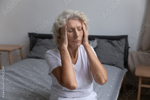 Unhappy unhealthy mature woman touching temples, suffering from strong headache or migraine, exhausted tired middle aged female feeling unwell, suffering from insomnia, sitting on bed at home