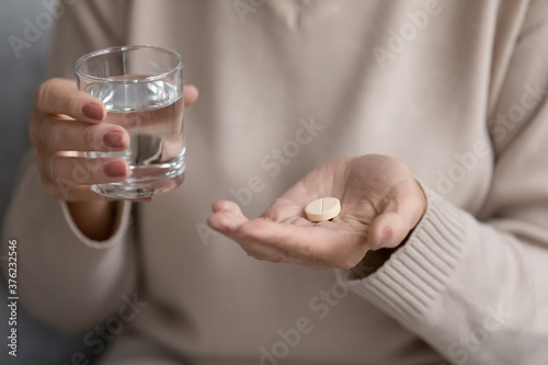 Close up mature woman holding glass of water and white round pill, middle aged female taking antibiotic or supplements, preparing to take emergency medicine, healthcare and treatment concept