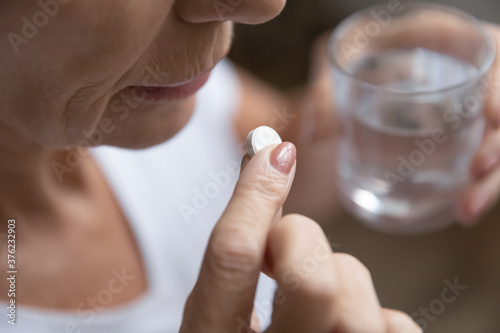 Close up senior mature woman taking painkiller  antibiotic  supplements or vitamin medication  middle aged female holding glass of water and white round pill in hands  disease treatment concept