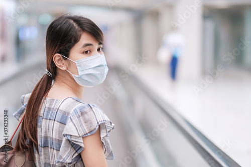 Young adult female wearing face mask in airport terminal, protection Coronavirus disease infection, Asian woman traveler walking on escalator in aerodrome. New Normal and travel under COVID-19 concept