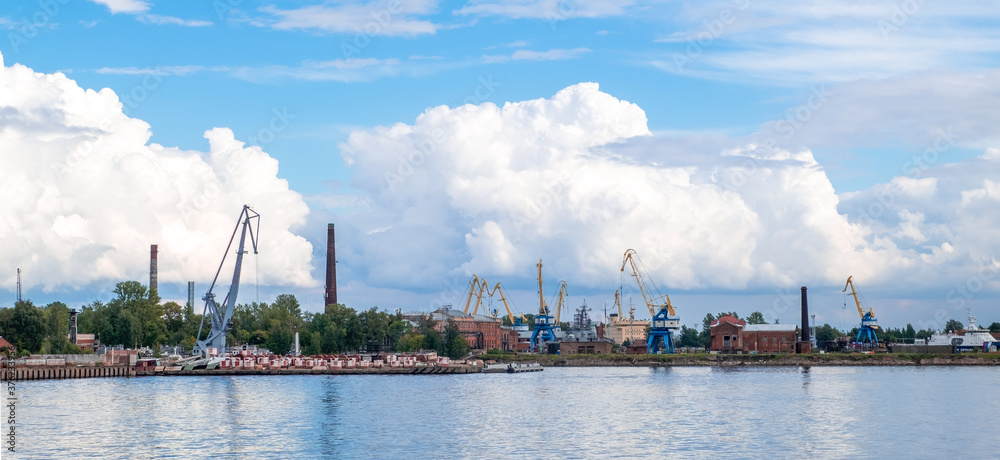 Kronstadt, St. Petersburg: August 29, 2020. View of the commercial port with quays, ships and cranes in loading area across the port water area against beautiful cumulus clouds. Selective focus.