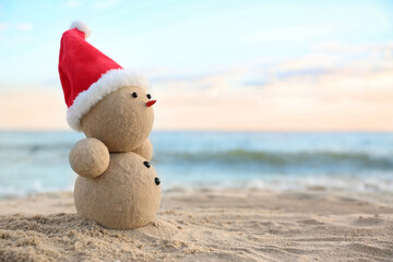 Snowman made of sand with Santa hat on beach near sea, space for text. Christmas vacation