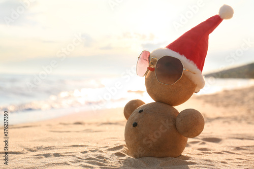 Photography Snowman made of sand with Santa hat and sunglasses on beach near sea, space for text