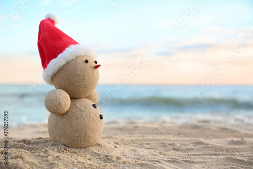 Fototapeta Naklejka Na Ścianę i Meble -  Snowman made of sand with Santa hat on beach near sea, space for text. Christmas vacation