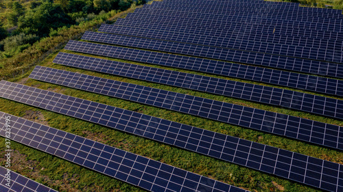 Solar Power Station in Green Field on Sunny day. Aerial view. Solar Panels Stands in a Row in Field for Power Production. Drone fly over Solar Farm. Renewable green energy. Alternative energy sources.