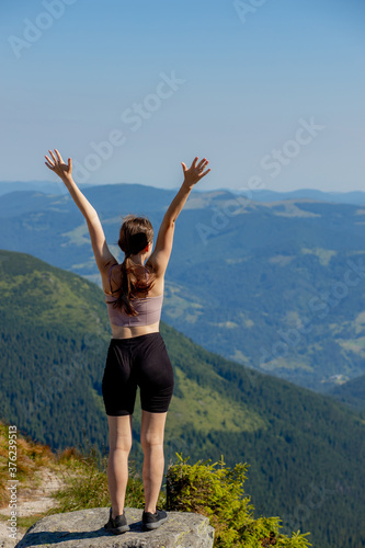 The young girl at the top of the mountain raised her hands up on blue sky background. The woman climbed to the top and enjoyed her success. Back view