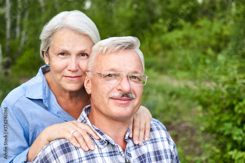 Elderly couple in love are sitting on a bench in the park, smiling and hugging against a background of green trees