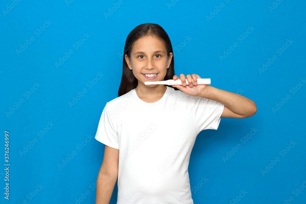 Brunette girl with long hair in a white T-shirt on a blue background brushes her teeth with an electric toothbrush . Brunette girl with long hair in a white T-shirt on a blue background brushes her teeth with an electric toothbrush .