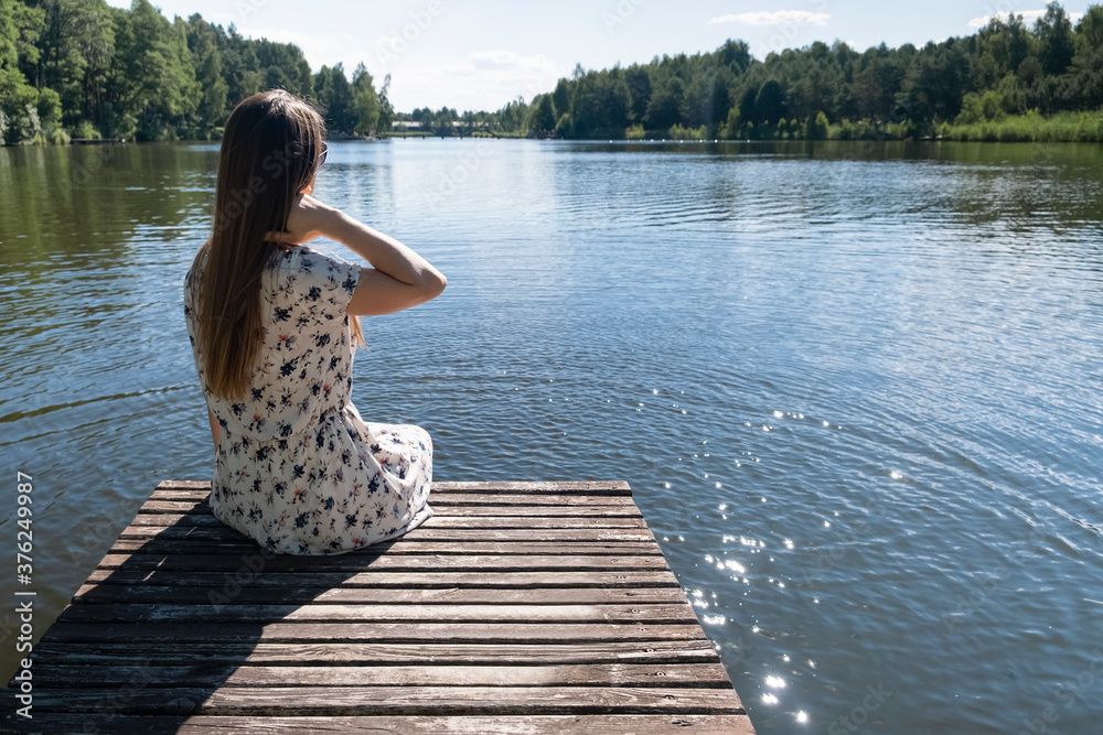 Naklejka premium Woman on the lake. Young woman wearing white dress enjoying nature and resting sitting on a wooden pier on the lake , back view