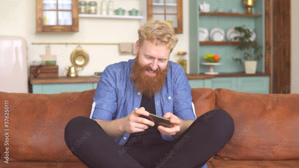 Ginger bearded man enjoy playing games on the smartphone sitting on couch at home Ginger bearded man enjoy playing games on the smartphone sitting on couch at home