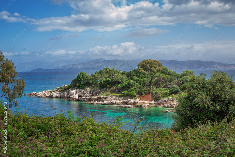 Beautiful landscape - sea lagoon with turquoise water, cliffs and rocks, covered with trees green grass and bushes, blue sky and mountains on the horizon. Corfu Island, Greece.