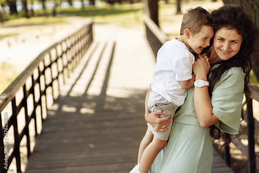 Fototapeta premium Portrait of adorable mother with overjoyed son at the park, beautiful woman hold in arms little boy, smiling, hugging, family time, motherhood and childhood concept