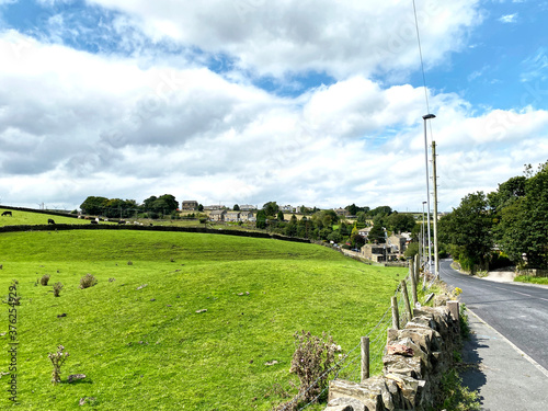 View, by a dry stone wall, of Brighouse Road, leading down into Denholme, with fields, houses and trees