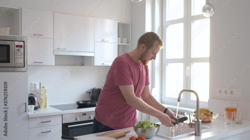 caucasian man washes vegetables for salad in a white light kitchen caucasian man washes vegetables for salad in a white light kitchen